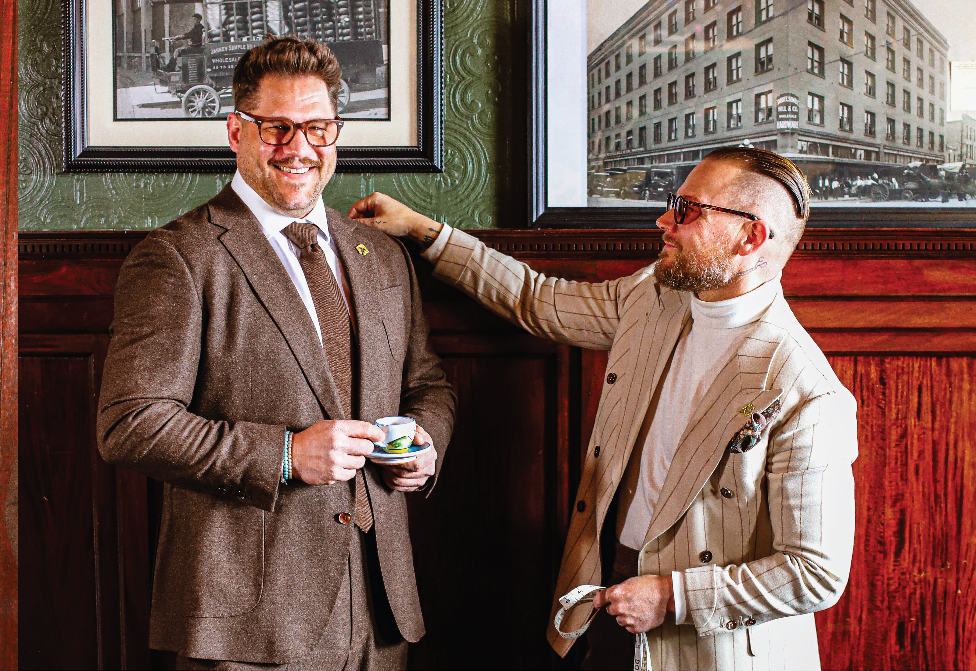 Two men in suits standing in a room with framed pictures on the wall.